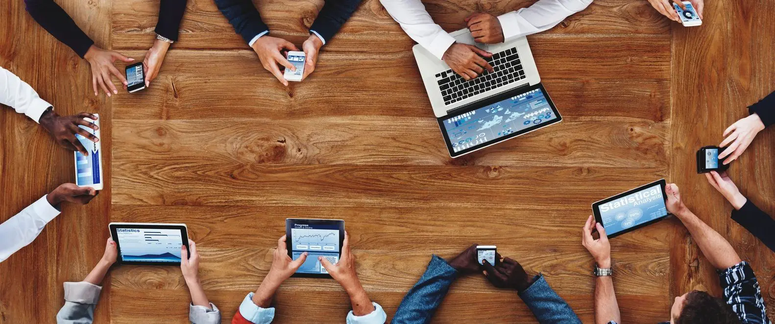 People using various devices—phone, tablet, laptop—at a wooden table.