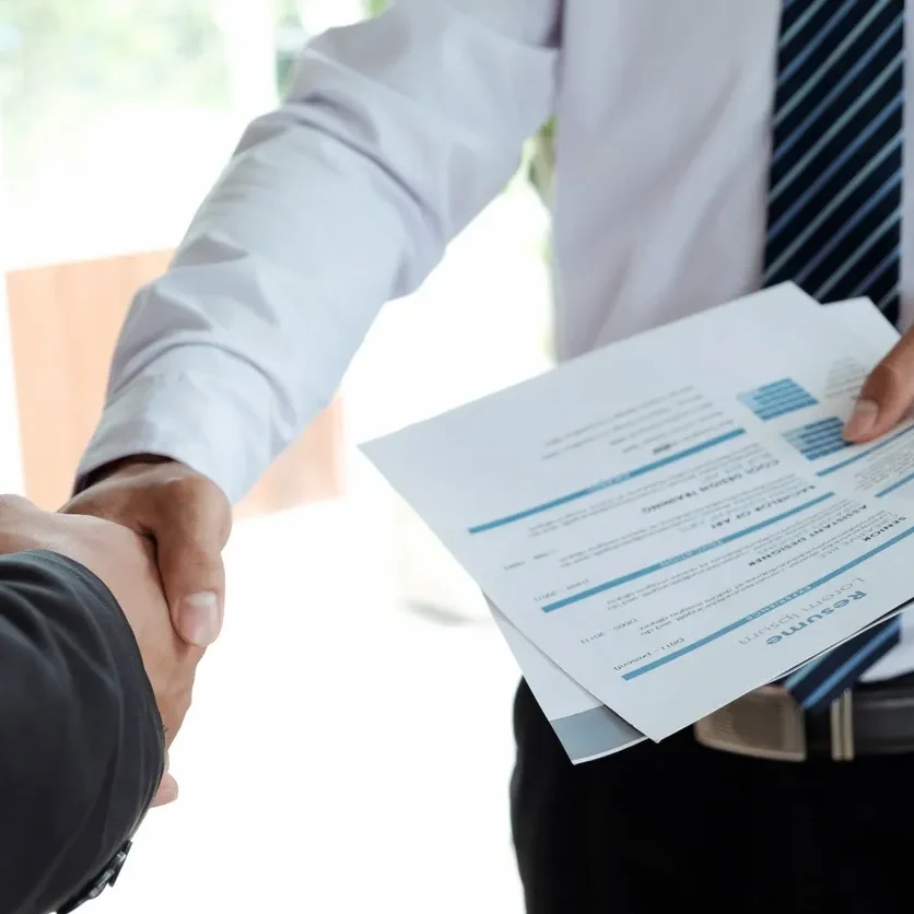 Two professionals shaking hands, one holding resumes.